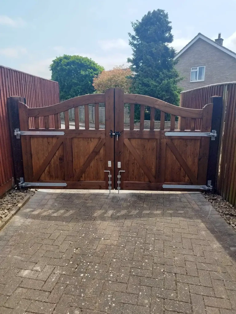 A wooden double gate with metal hinges and a central lockbar opens onto a brick-paved driveway. The gate is surrounded by wooden fencing, and in the background, there's a house and trees under a clear sky.