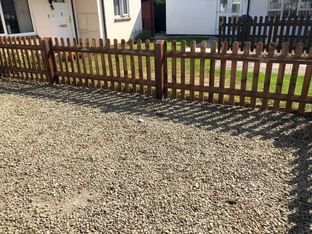 A gravel driveway leads to a wooden picket fence casting striped shadows. Behind the fence is a grassy area and houses with white walls and windows. Sunlight enhances the contrast between the shadows and ground.