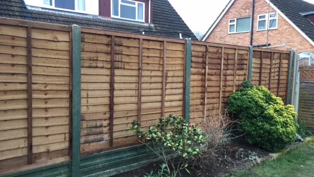 A wooden fence in a backyard garden with green and leafy shrubs in front. Two houses with slanted roofs are visible behind the fence. The sky is overcast.