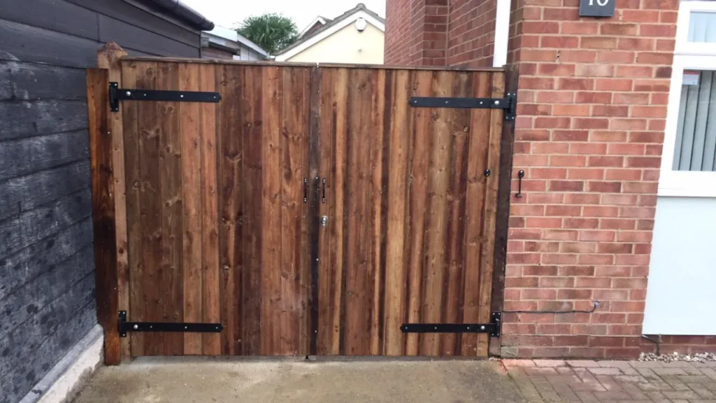 A wooden gate with metal hinges and a latch, set between a brick wall and a wooden fence, leading to a paved pathway. The gate is made of vertical wooden planks with a natural finish. A portion of a house exterior is visible.