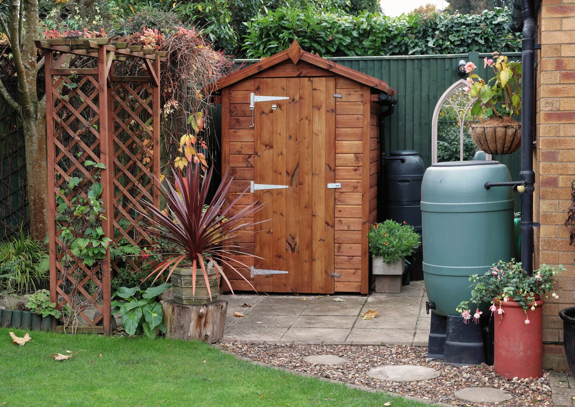 A tidy garden features a wooden shed with a pitched roof, surrounded by various plants and flowers. A trellis and large green water butt are nearby. The shed is on a patio with a lush lawn in the foreground.