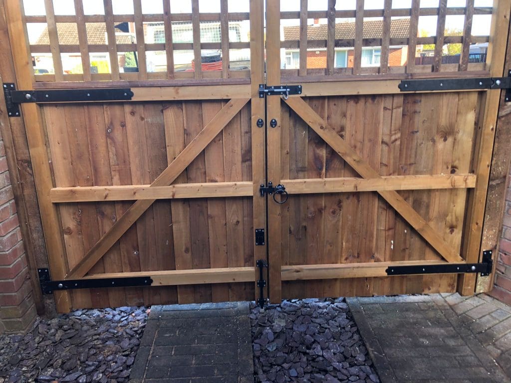 A wooden gate with a cross-brace design is secured with black metal hinges and a latch. It leads to a paved area with houses and rooftops visible beyond. The ground in front of the gate has dark stones and brick paving.