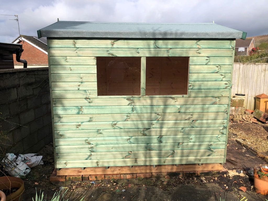 A small, light green wooden shed with a flat roof and two rectangular windows stands in a backyard. It's built on a base of wooden blocks, and garden soil bags are visible nearby. The sky is partly cloudy.