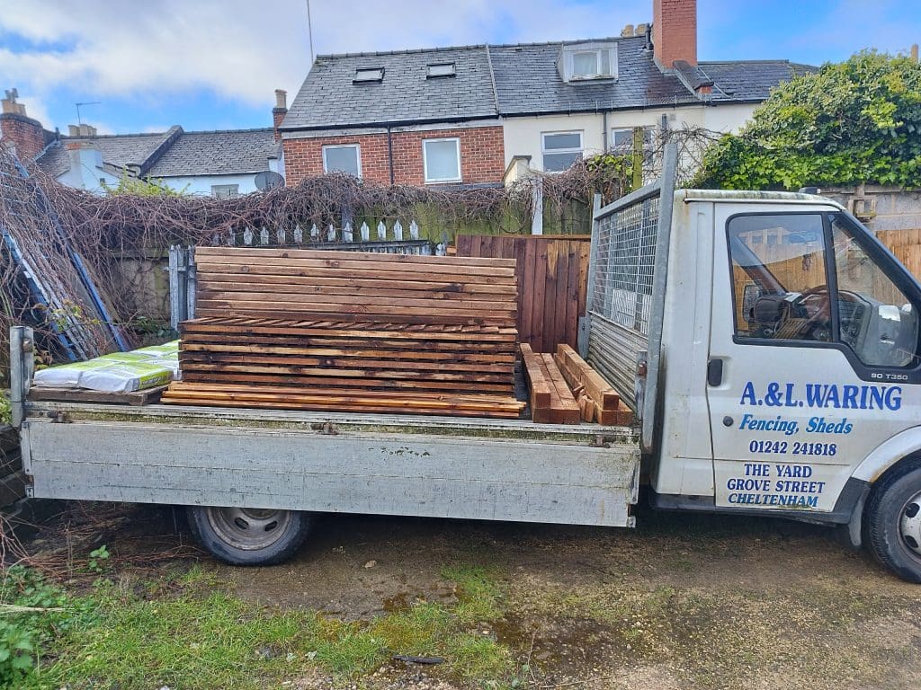 A flatbed truck loaded with stacked wooden planks is parked in a yard. The vehicle has text on the side with contact information. In the background, there are residential buildings and a cloudy sky.