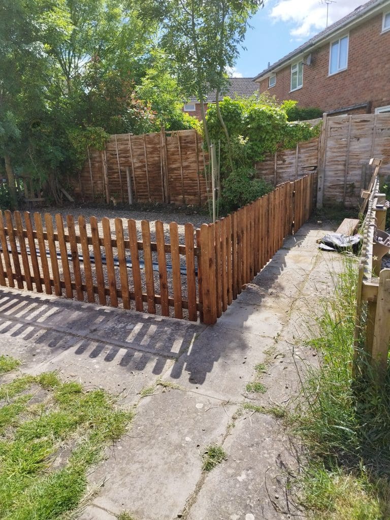 A backyard with a wooden fence surrounding a garden area. The garden is mostly bare soil, bordered by tall wooden panels and lush green trees. A brick building with white-trimmed windows is visible in the background.