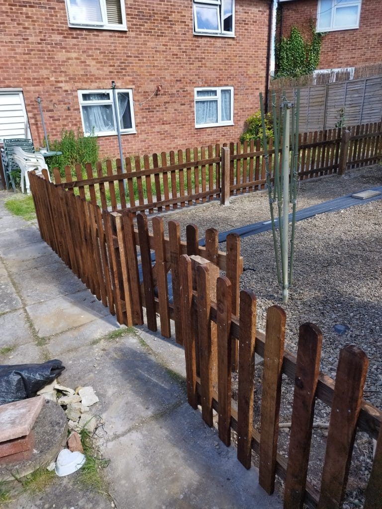 A wooden picket fence encloses a gravel yard next to a brick building with white-framed windows. A green pole stands inside the fenced area. A path leads up to the gate of the fenced section.