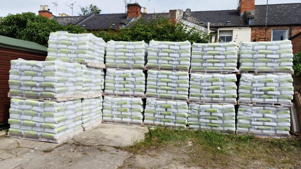 Stacks of white and green bags piled on wooden pallets, arranged in rows in a yard. Residential buildings and trees are visible in the background.