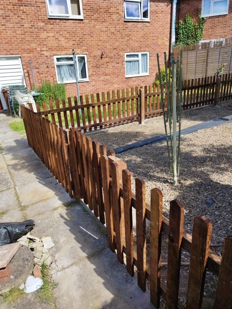 A wooden picket fence encloses a gravel area with a narrow path. A tall, thin plant grows inside the fenced area. Red brick buildings are in the background with visible windows and doors.