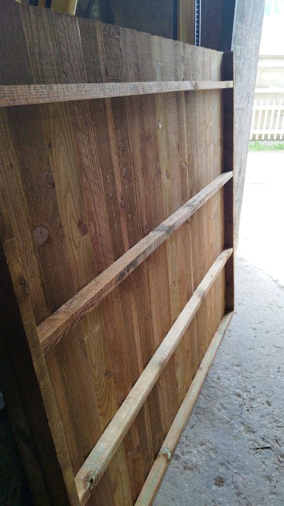 Wooden fence panel with horizontal support beams leaning against a wall in a well-lit area, possibly a workshop or garage. Sunlight filters in from the right, illuminating the wood grain and texture. A white picket fence is visible in the background.