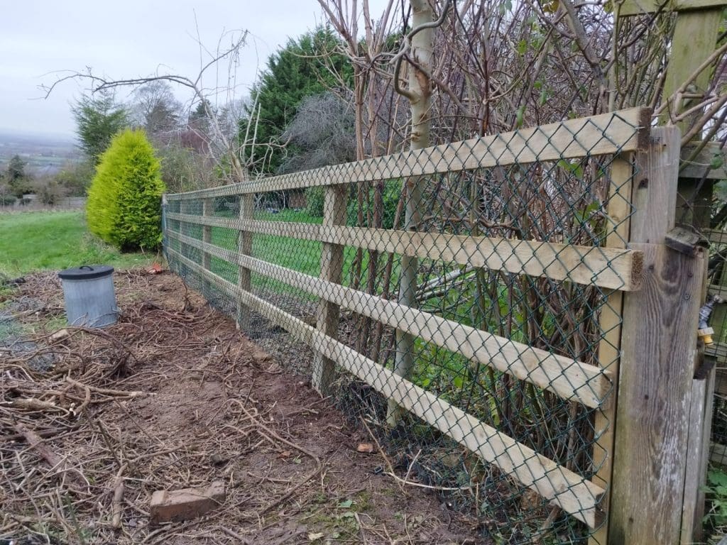 A wooden and wire fence runs along a garden, with bare trees and bushes beside it. A distant view of fields and hills is visible in the background, under an overcast sky. A plastic bin is on the ground near the fence.