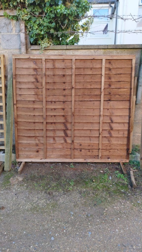 A wooden fence panel stands upright against a wall. The panel consists of horizontal slats supported by vertical and horizontal framing. Ivy grows over the wall above, and there is a dirt and gravel ground surface in front.