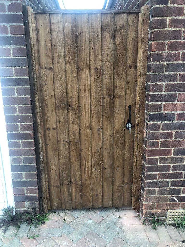 A wooden gate with vertical panels is framed by two brick pillars. It has a black metal handle and a latch. The pathway leading to the gate is paved with herringbone-patterned bricks.