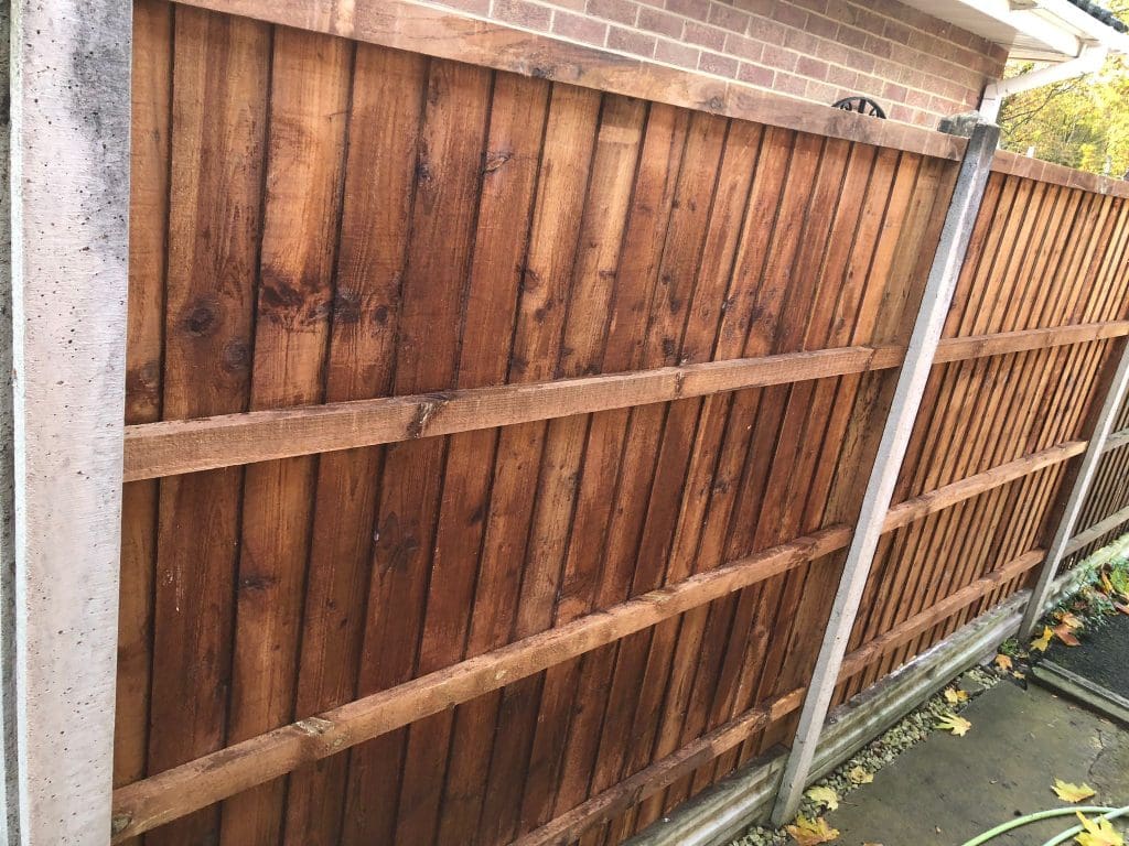 A tall wooden fence with vertical slats and horizontal support beams stands between concrete posts. The fence is next to a brick building and the ground is covered with some leaves and a garden hose.