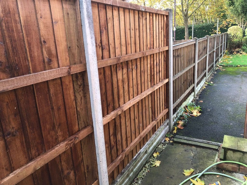 A perspective view of a long wooden fence with vertical panels, supported by concrete posts. The ground is lined with fallen leaves and a garden hose is coiled in the foreground. Trees and a path run alongside the fence in the background.