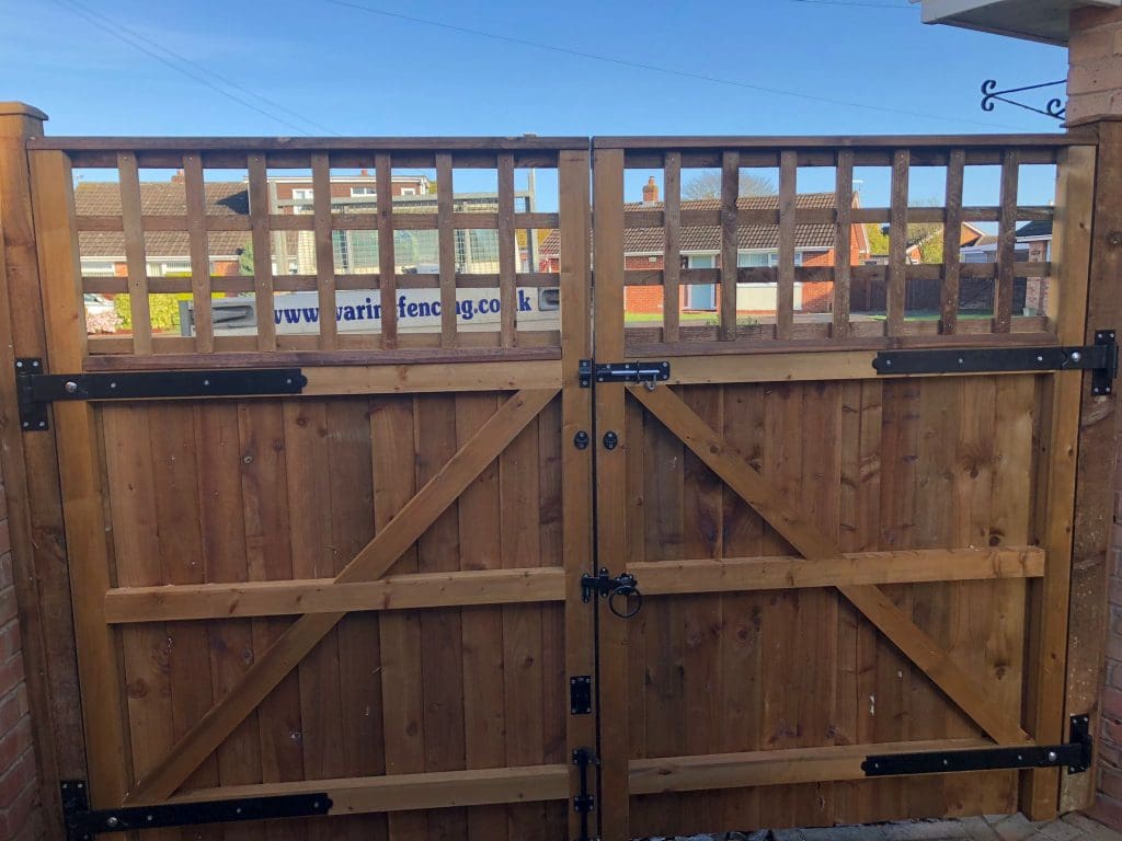 A wooden double gate with a lattice top and metal hinges and latch is closed, flanked by brick pillars. In the background, there are residential buildings and a partially visible sign with text. The sky is clear and blue.