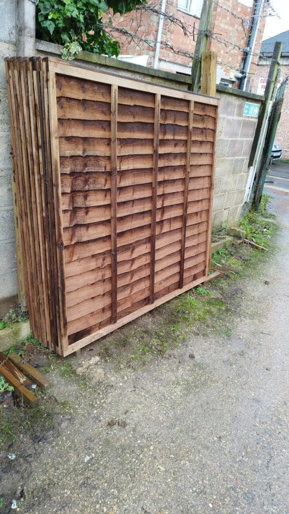 A stack of wooden fence panels leaning against a concrete wall in an outdoor setting, near a narrow path. The panels have a brown finish and are set on a slightly uneven ground with some grass and mud.