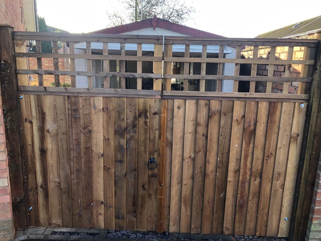 A wooden gate with a lattice design on top stands between two brick walls. Behind the gate, a small shed or garage with a red roof is visible. The ground is covered with gray stones.