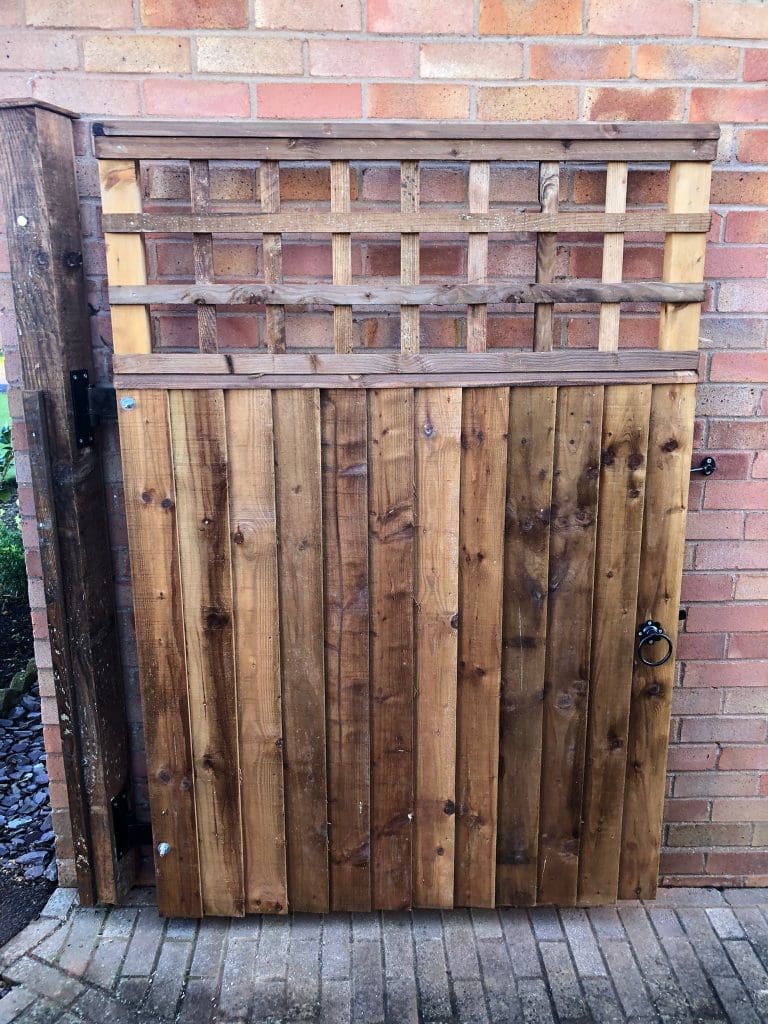 A wooden gate with a lattice design on top, set against a brick wall. The gate features a dark metal handle and hinges, providing access to a garden or courtyard. The wood is stained a natural brown, contrasting with the light-colored bricks.