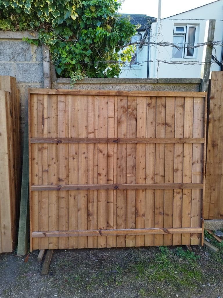 A wooden fence panel leaning against a concrete wall, with green ivy growing on one side. Barbed wire is visible on top. Other fence panels are nearby, and a building with a window is in the background.