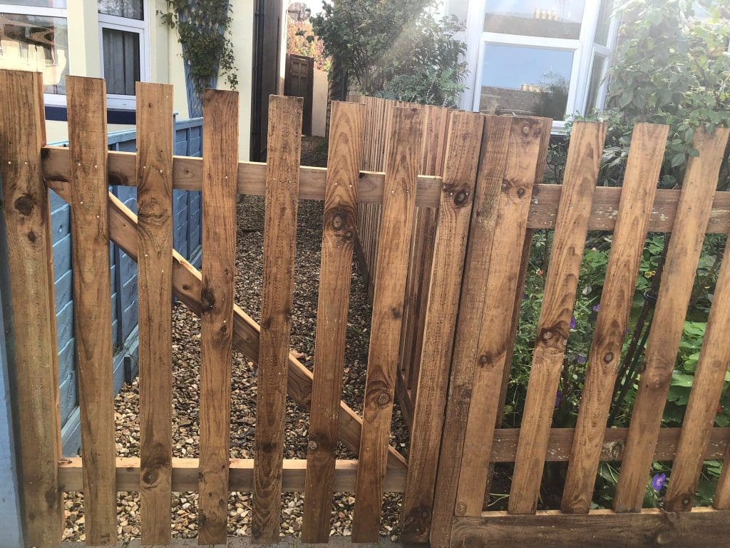 A wooden gate with a latch stands between fence panels, leading to a gravel pathway. The path is flanked by a blue building on the left and greenery on the right. Sunlight filters through, casting soft shadows on the ground.