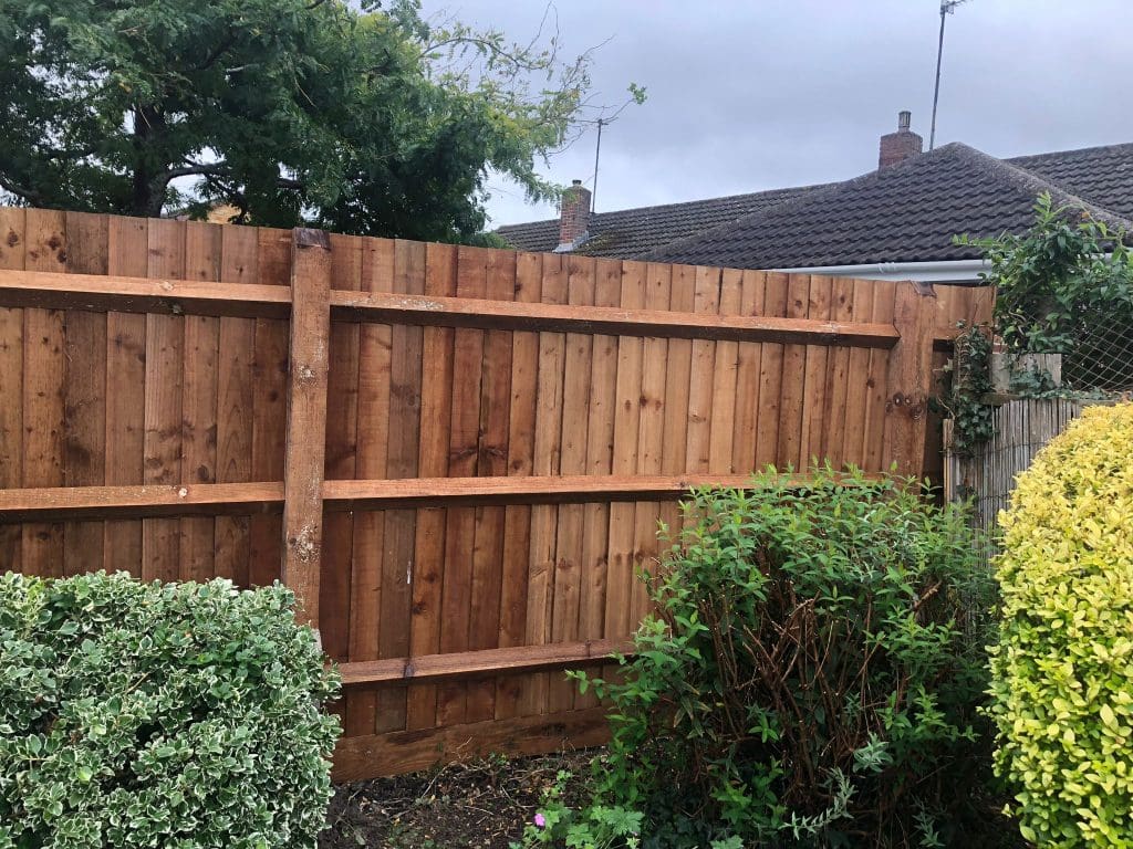 A wooden fence with horizontal support beams stands in a garden. Various shrubs and green plants grow in front of the fence. A cloudy sky and some single-story houses with brick chimneys are visible in the background.