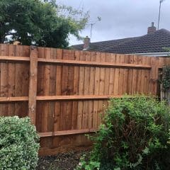 A wooden fence with horizontal support beams stands in a garden. Various shrubs and green plants grow in front of the fence. A cloudy sky and some single-story houses with brick chimneys are visible in the background.