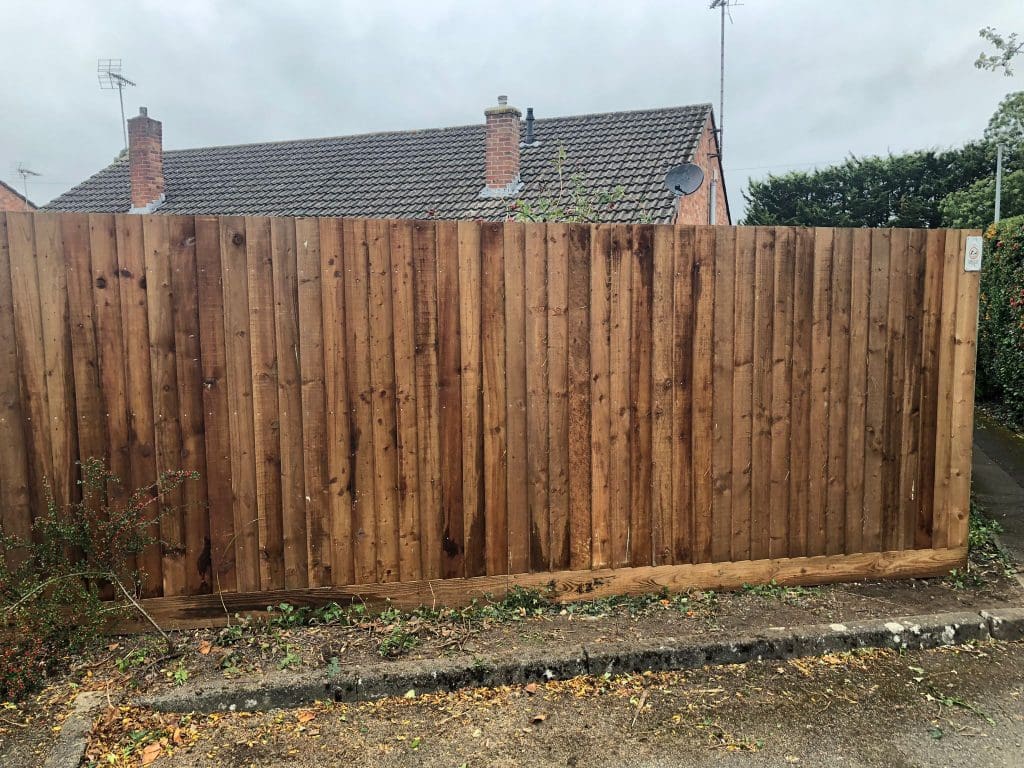 A tall wooden fence made of vertical planks, set in front of a suburban house with a tiled roof. The sky is overcast, and there's a patch of greenery at the bottom left corner.