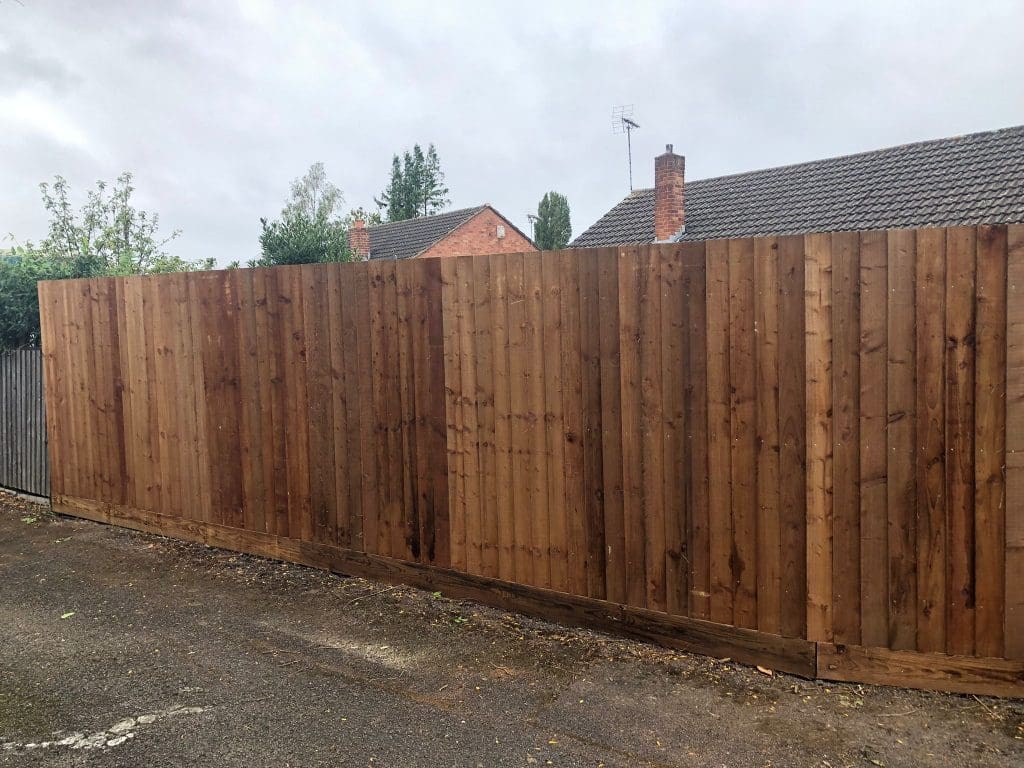 A tall, wooden fence with vertical planks stretches along a paved alleyway. In the background, residential houses with red brick and grey roofs are partially visible under an overcast sky.