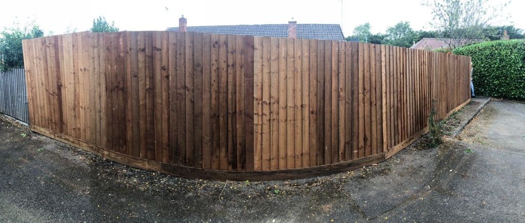 A tall wooden fence with vertical slats encloses a backyard. The fence is weathered and bordered by a paved driveway and greenery. Houses and trees are visible in the background under a cloudy sky.