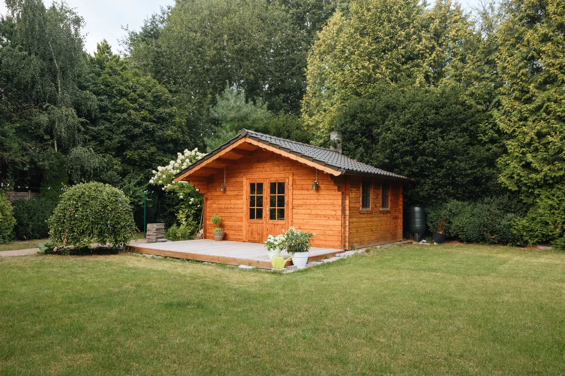 A small wooden cabin with a front porch sits amidst lush greenery. Surrounding the cabin are trees and bushes, and a well-kept lawn stretches out in front. Potted plants adorn the porch, adding to the rustic charm of the scene.