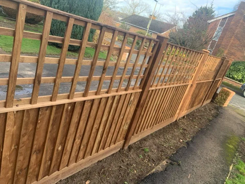 A newly constructed wooden fence with vertical slats and a grid pattern on top lines the edge of a garden path. Behind the fence, a road and residential houses are visible, along with some trees and a grassy area.