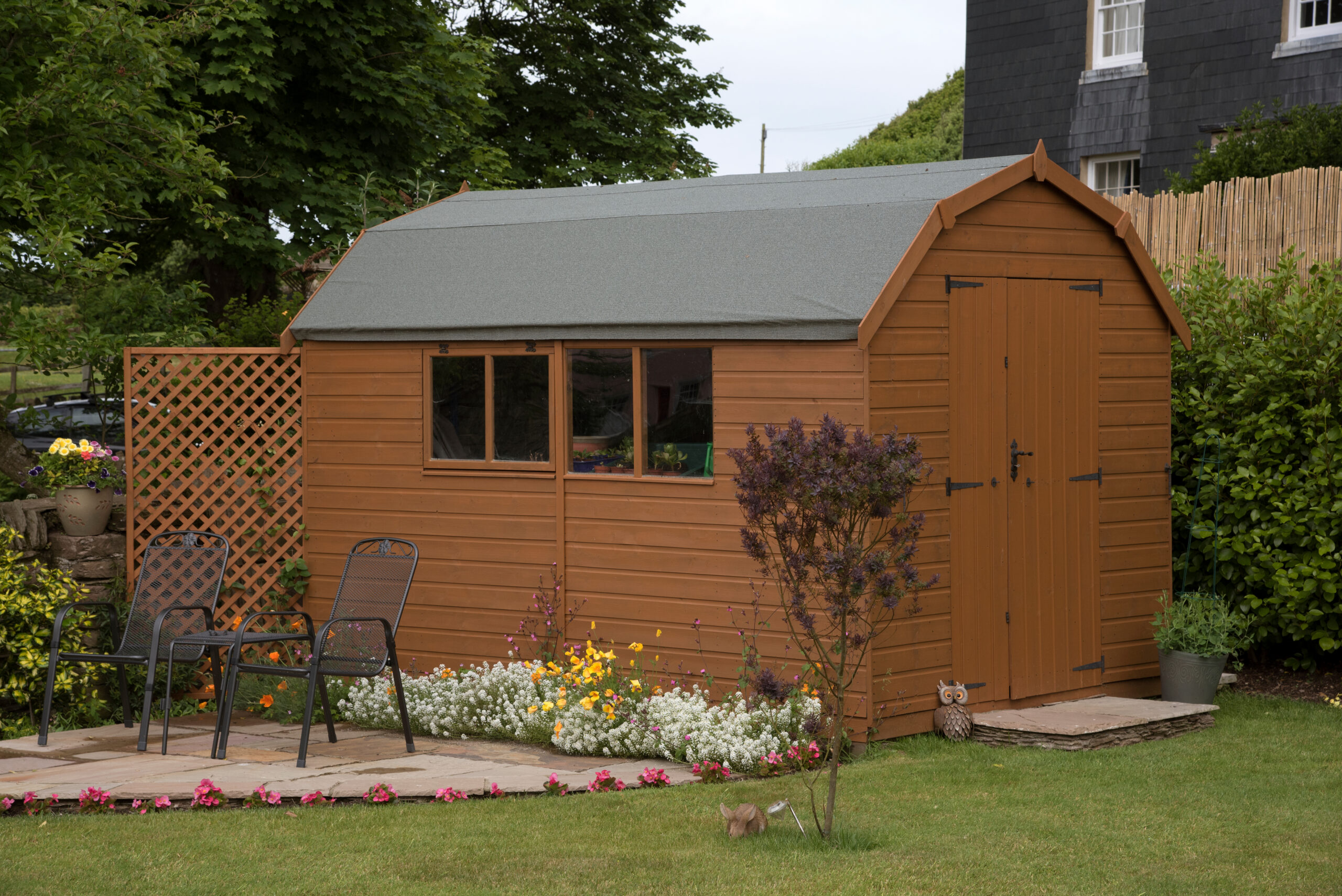 A wooden garden shed with a gray roof stands beside a small patio area with two black metal chairs and a table, surrounded by flowers and greenery in a landscaped yard.