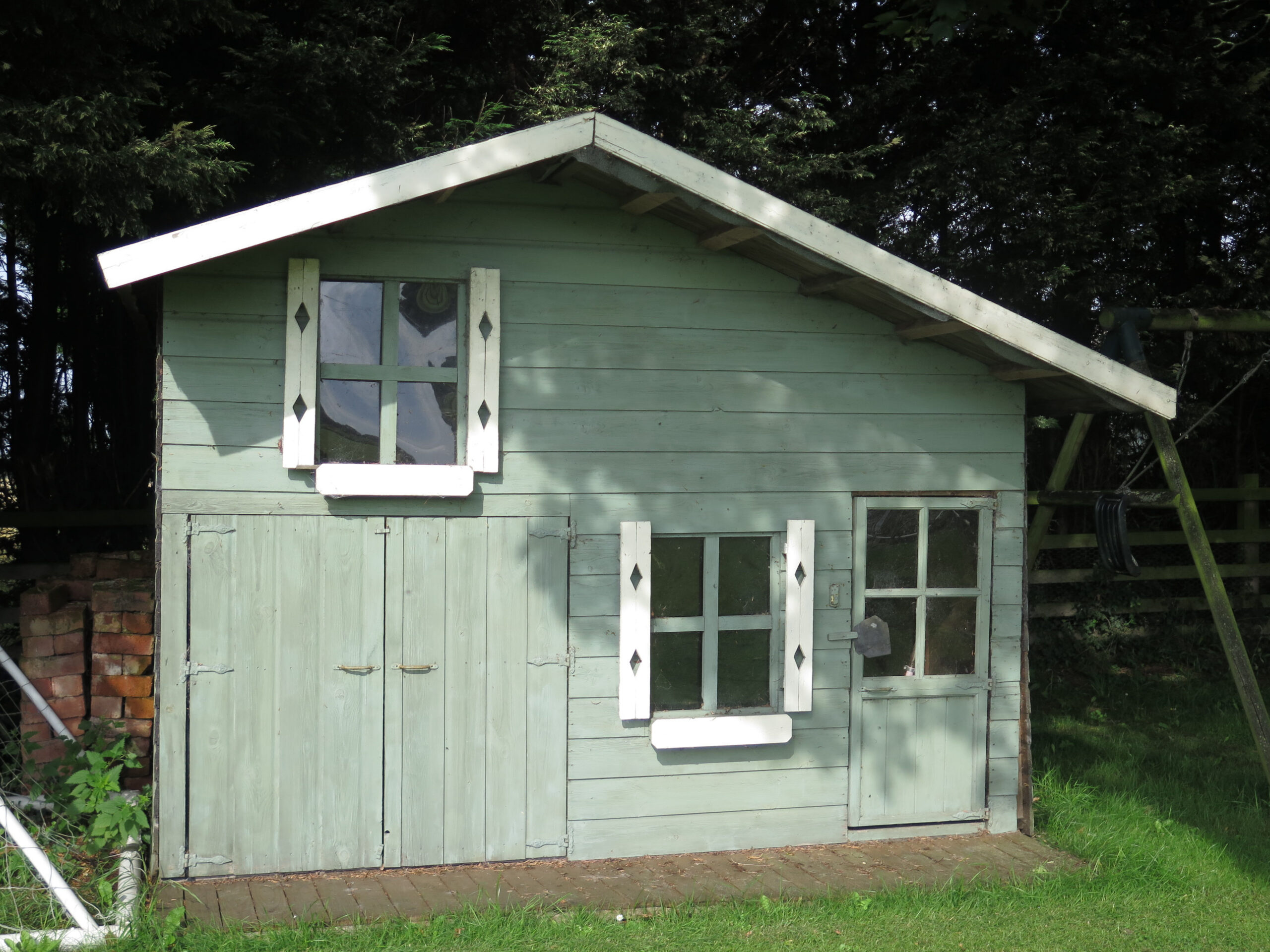 A small, pale green wooden playhouse with a pitched roof, white trim, and decorative window shutters, standing on a grassy lawn with trees and a brick wall in the background.