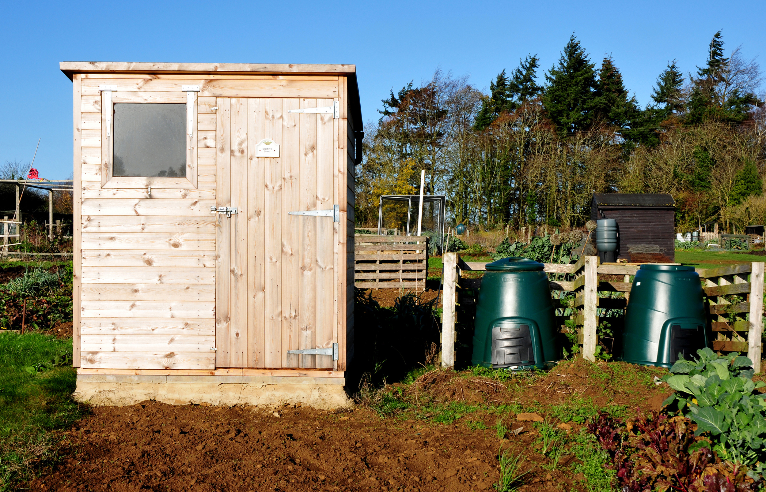 A wooden garden shed stands next to two green compost bins in a sunny allotment with trees and a smaller shed in the background. The soil and plants are visible around the shed.