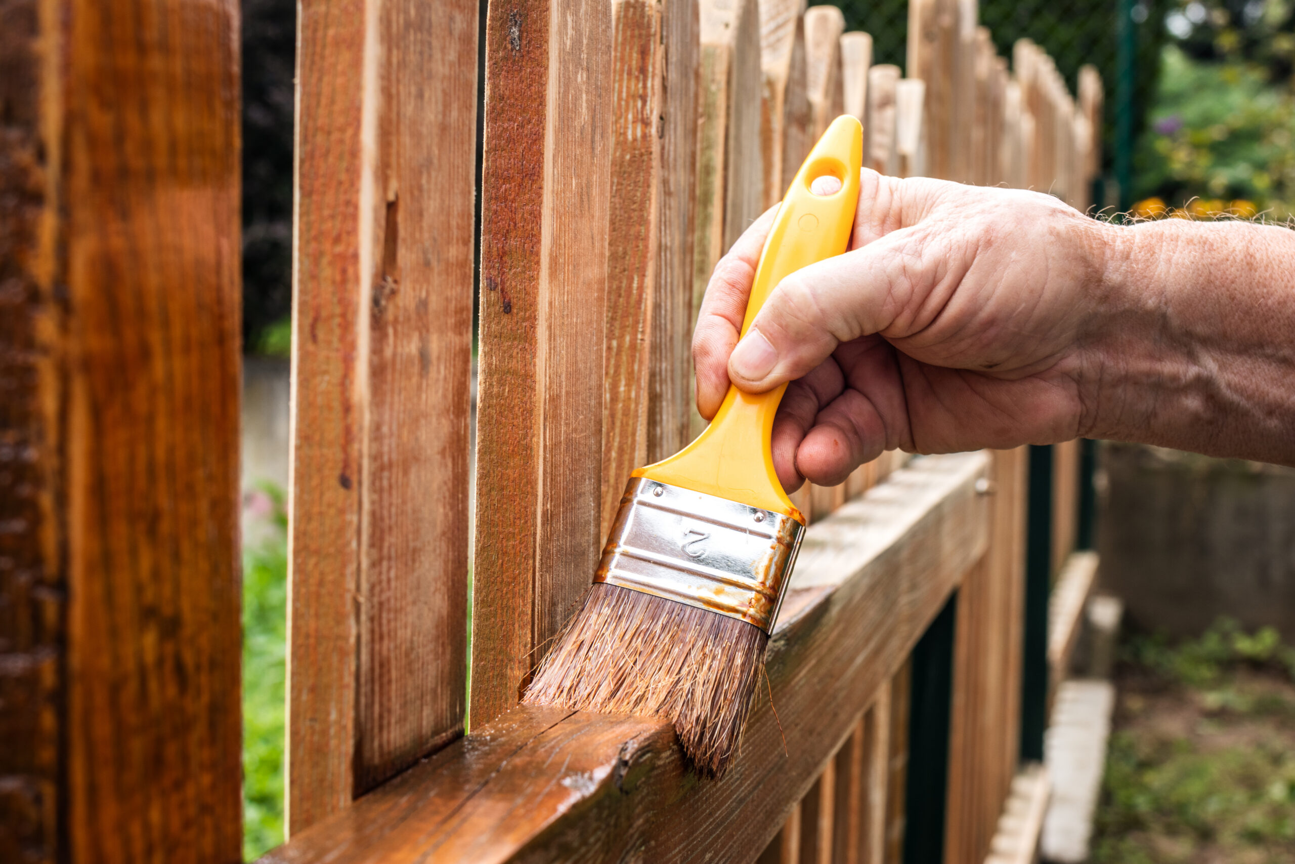 A person’s hand is painting a wooden fence with a yellow-handled paintbrush, applying a fresh coat of brown stain to the wood outdoors.