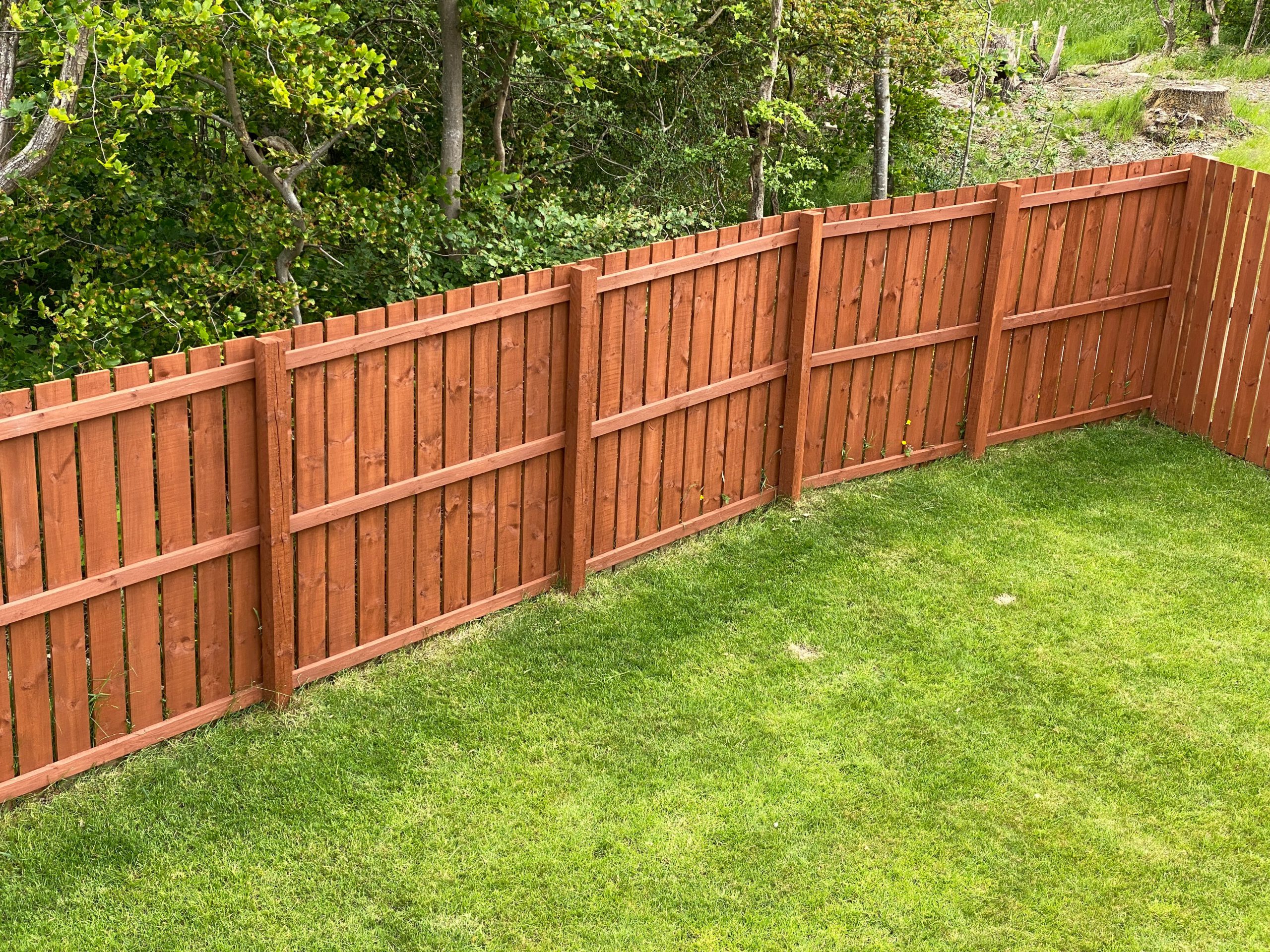 A wooden fence with vertical slats and horizontal support beams borders a neatly mowed green lawn, with trees and bushes in the background.