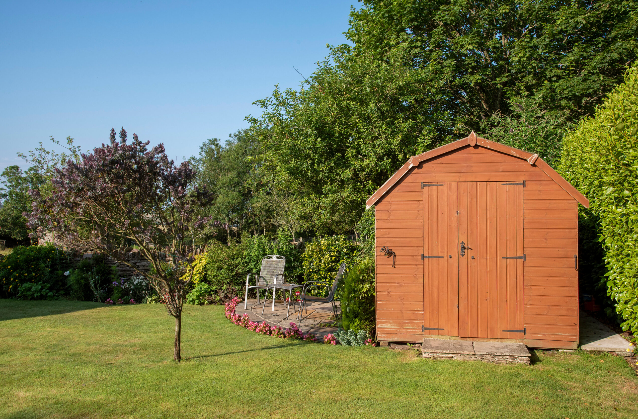 A wooden garden shed sits on a neatly mowed lawn, surrounded by lush greenery, flowering plants, and trees. A small patio with two chairs and a table is nearby under clear blue sky.