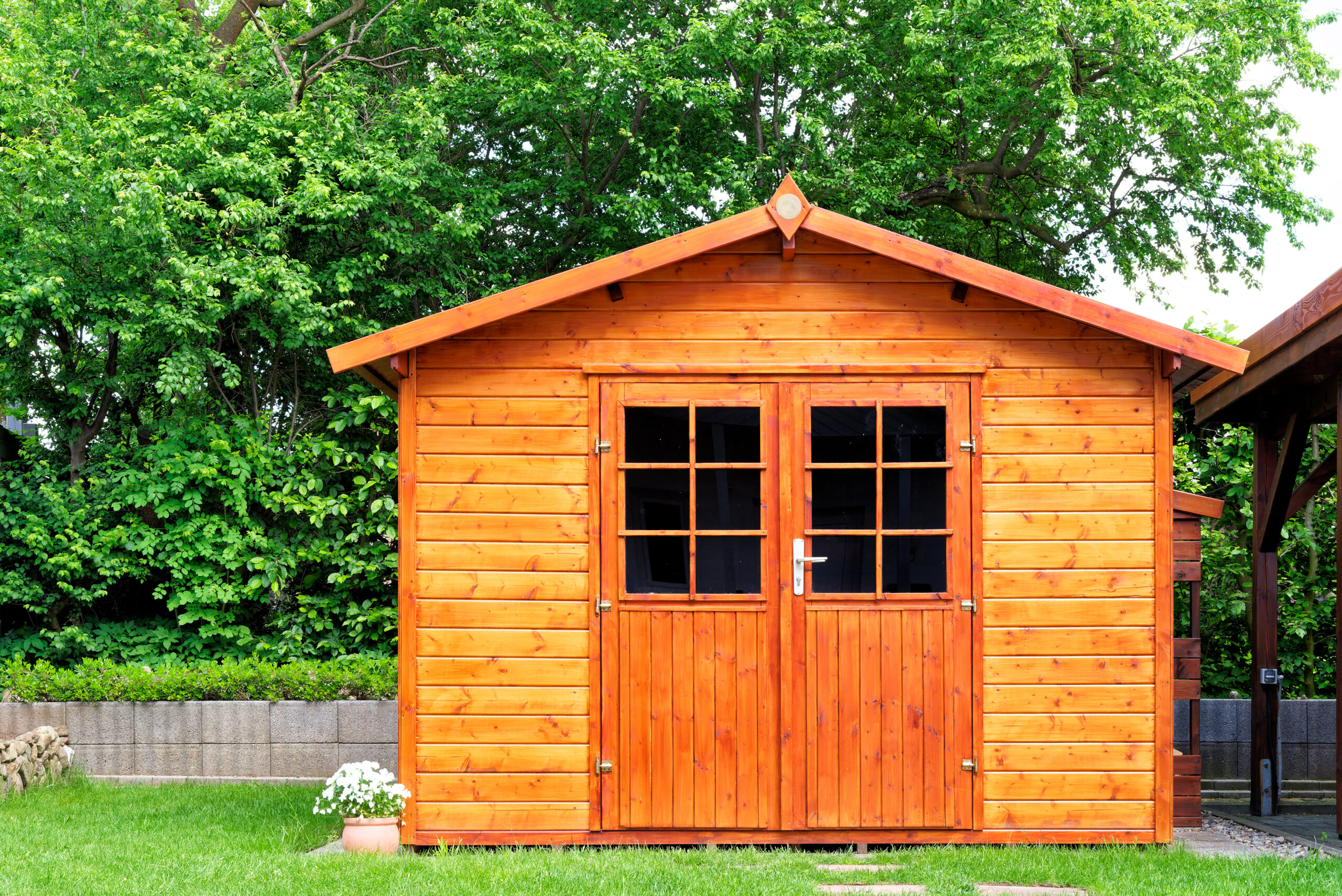 A wooden garden shed with double doors and small windows stands on green grass, surrounded by lush trees and plants.
