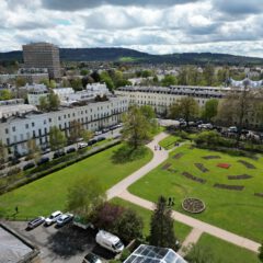 Aerial view of a city park with landscaped flower beds, pathways, and surrounding white terraced buildings; cars are parked along the street and distant hills are visible under a partly cloudy sky.
