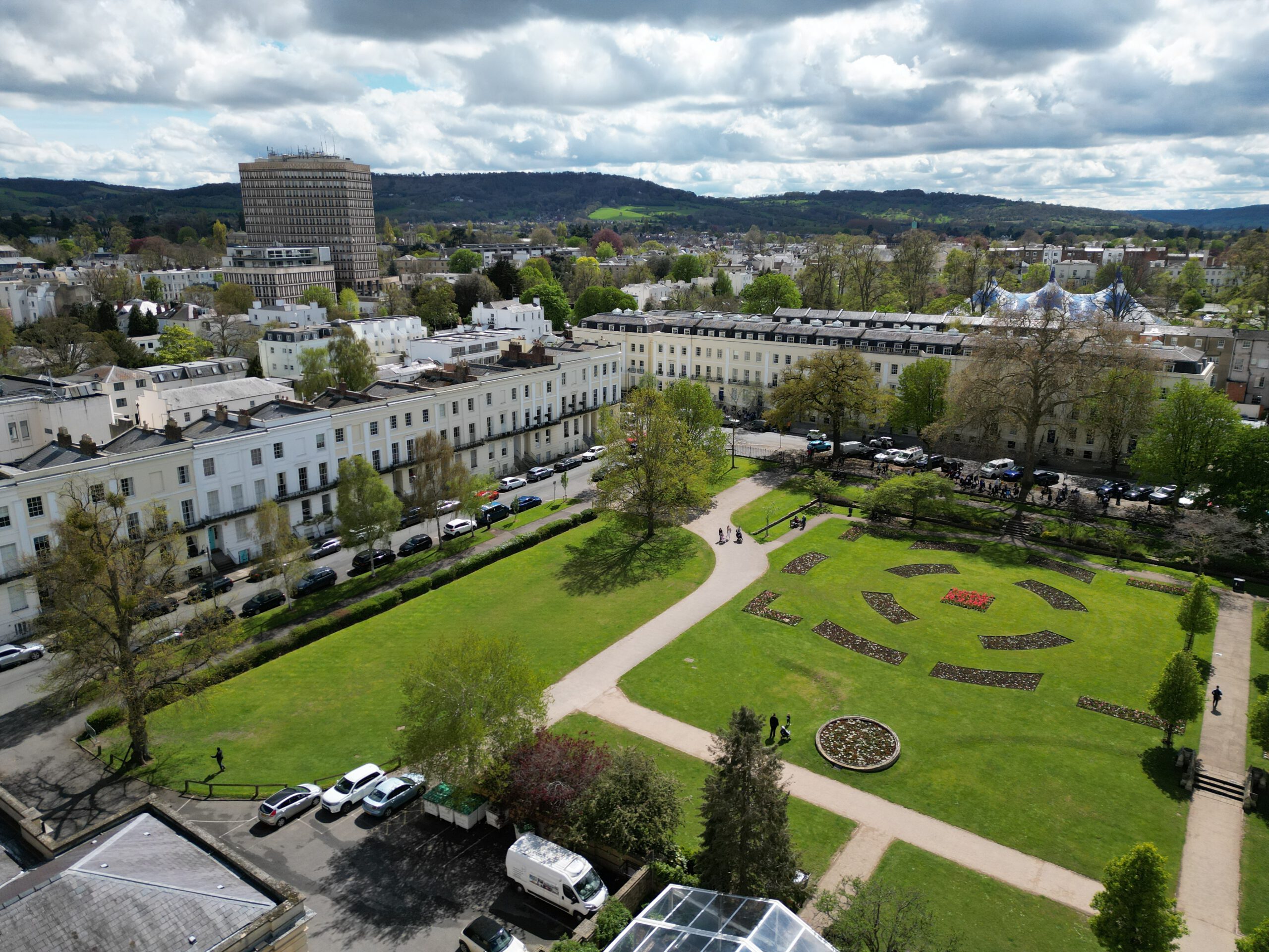 Aerial view of a city park with landscaped flower beds, pathways, and surrounding white terraced buildings; cars are parked along the street and distant hills are visible under a partly cloudy sky.