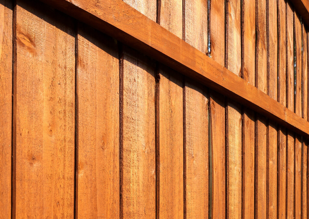 Close-up view of a wooden fence with vertical planks and a horizontal beam, all stained in a warm, golden-brown color. The wood grain and texture are clearly visible in the sunlight.