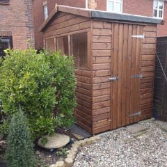 A wooden garden shed with a window and a closed door sits beside a brick house, surrounded by green bushes, small plants, and a gravel path.