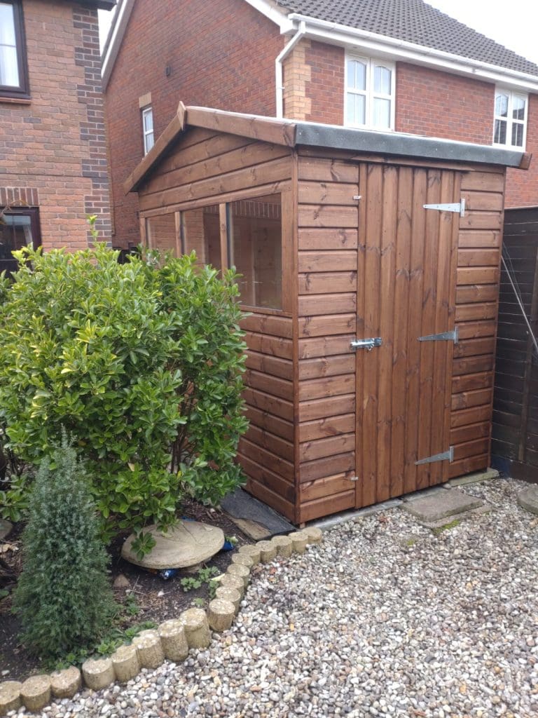 A wooden garden shed with a window and a closed door sits beside a brick house, surrounded by green bushes, small plants, and a gravel path.