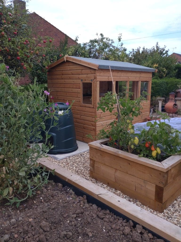 A wooden garden shed sits in a backyard surrounded by plants, a black compost bin, a raised wooden planter with vegetables, gravel paths, and a brick outdoor fireplace. Trees and shrubs fill the background.