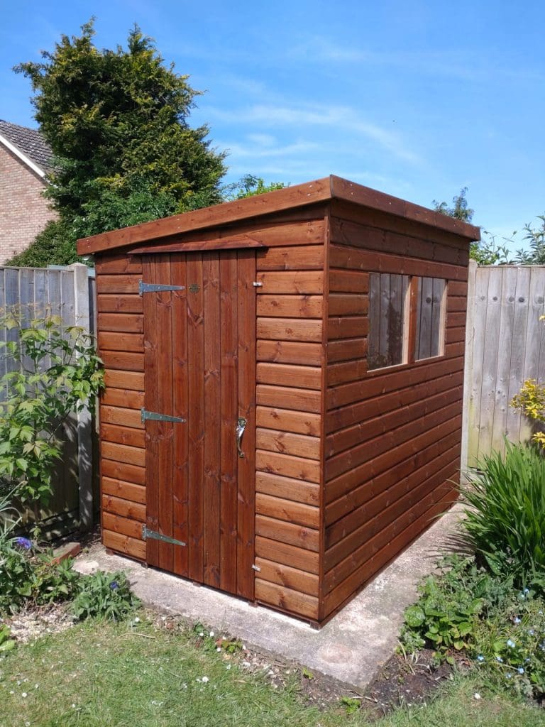 A wooden garden shed with a sloped roof, window on one side, and a closed door, standing on a concrete base in a fenced backyard with grass and plants around it.