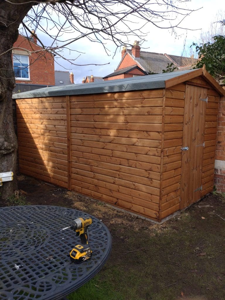 A wooden garden shed with a slanted roof stands beside a tree and a brick wall. A round metal table with a drill and screws is in the foreground, and houses are visible in the background.