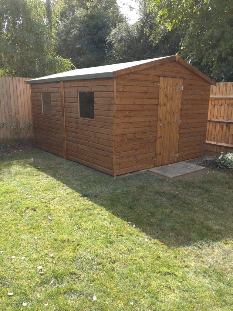 A wooden garden shed with a pitched roof and two windows stands on a grassy lawn, surrounded by a wooden fence and bordered by trees and plants. Sunlight casts shadows on the shed and lawn.