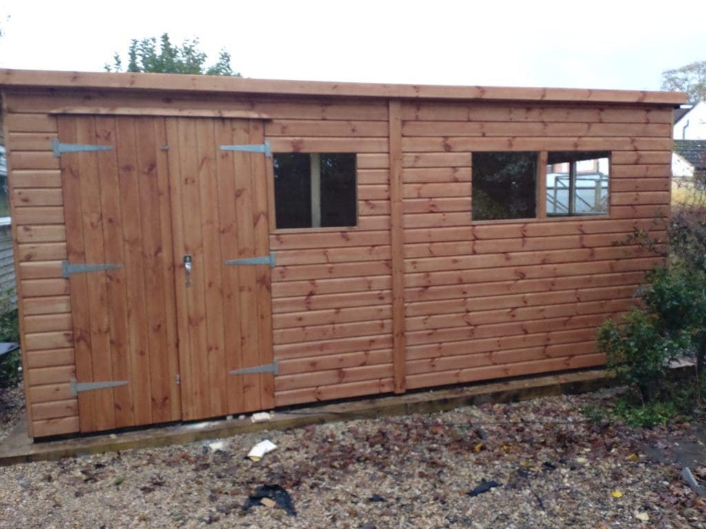 A large wooden garden shed with two windows and a single door, set on a gravel area with some plants and fallen leaves in front. The shed has horizontal wooden panels and metal hinges on the door.