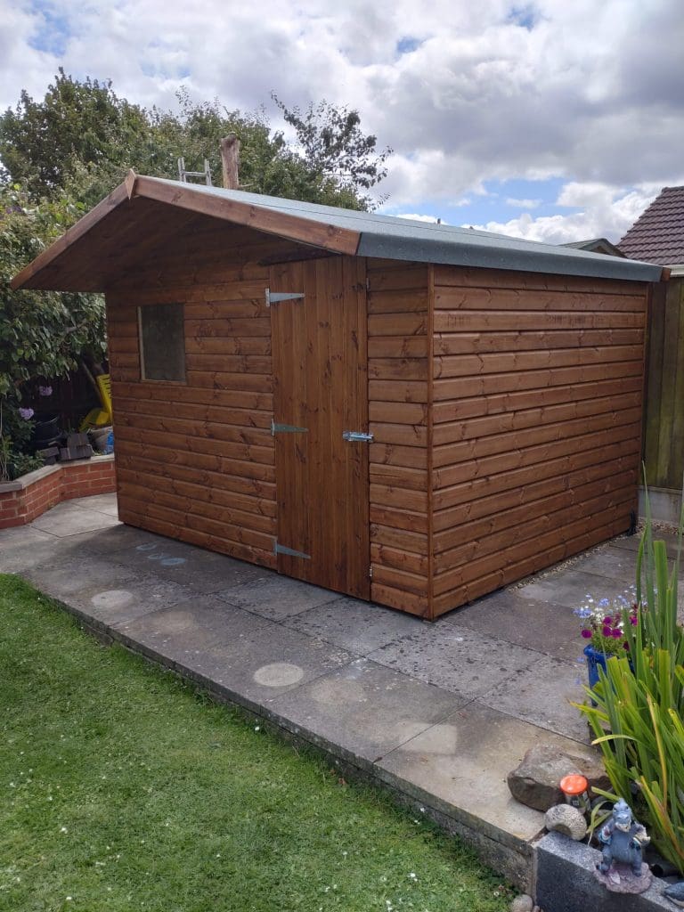 A wooden shed with a pitched roof and a small window stands on a paved area in a garden, surrounded by grass, plants, and a few garden ornaments under a partly cloudy sky.