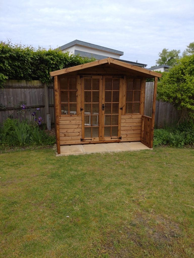 A small wooden garden shed with double glass doors, situated on a lawn in front of a wooden fence and surrounded by greenery and plants.